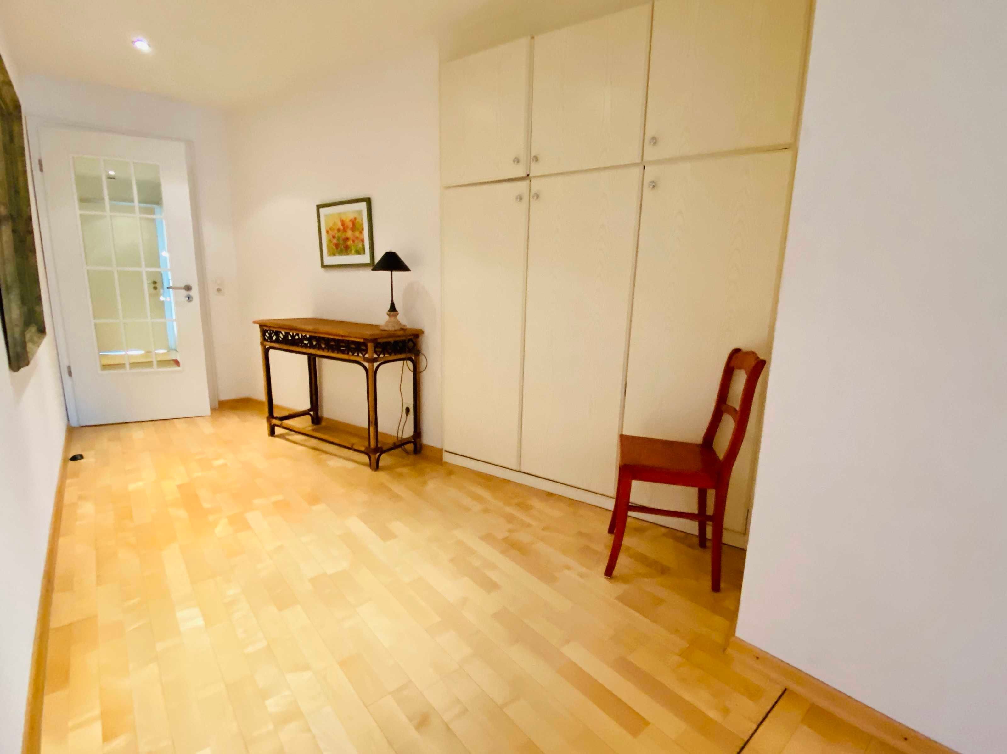 A room with a wooden floor, a table with a lamp, a chair, and a wardrobe with multiple doors.