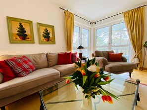 A living room with two sofas, a glass coffee table, and a vase with flowers. There are two windows with curtains, and a painting on the wall.
