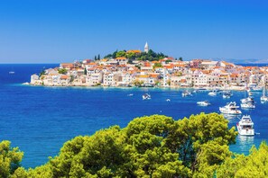 Une ville côtière qui se distingue par une église imposante située sur une colline, une mer d’un bleu limpide et de nombreux bateaux ancrés à proximité.