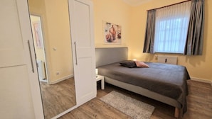 Et soveværelse med seng, natbord, radiator, dør og et vindue med gardiner.
