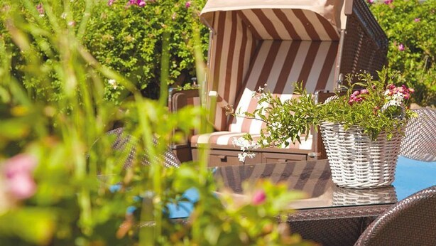 Une chaise en osier surmontée d’un parasol à rayures, une table en osier et un pot de fleurs avec des fleurs roses.