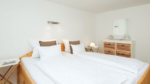 Une chambre avec un lit en bois, du linge de lit blanc, une table de chevet avec une lampe, une petite table avec un livre et un miroir sur le mur.