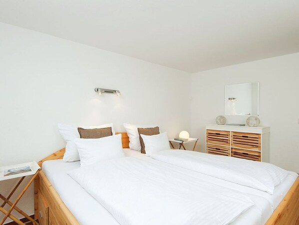 A bedroom with a wooden bed, white bedding, a bedside table with a lamp, a small table with a book, and a mirror on the wall.