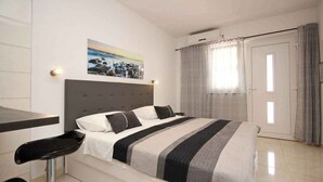 Un dormitorio con cama, mesita de noche, una silla, un cuadro en la pared y una puerta con cortinas.