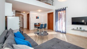 Un amplio salón con sofá, una mesa de comedor con sillas, televisor montado en la pared y un balcón con barandilla.