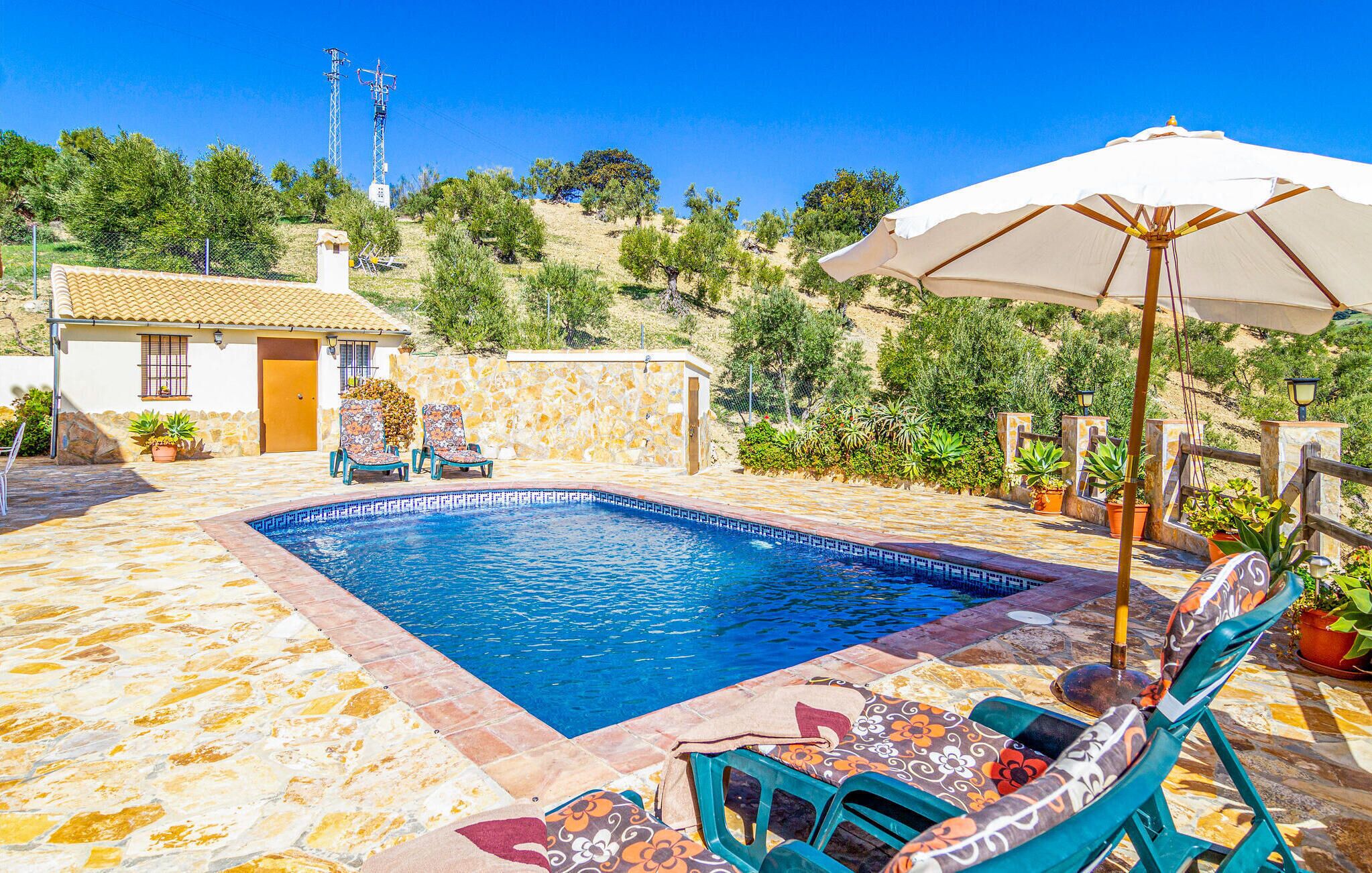 Una piscina con piso de baldosas, dos sillas de descanso con cojines de colores y una sombrilla grande.