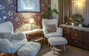 Ein gemütliches Zimmer mit floralem Wandpapier, einer Weltkarte, einer Lampe, weichen Sesseln, einem Holznachttisch und einer Kommode mit Dekorationsobjekten.