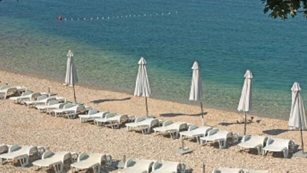 Una playa con hileras de tumbonas y sombrillas, un mar azul cristalino y una orilla pedregosa.