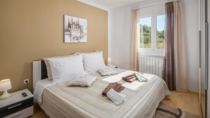 Ein ordentlich bezogenes Bett mit weißen Laken und einer beigen Decke, ein Nachttisch mit einer Lampe und ein Gemälde an der Wand.