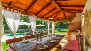 Ein Essbereich im Freien mit einem Holztisch, gedeckt für eine Mahlzeit, einem Backofen aus Ziegelsteinen und einem Pool im Hintergrund.