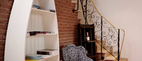 Een kamer met een witte, gebogen boekenkast, een gestreept fauteuil, een houten bijzettafeltje, een klein houten kastje, en een trap met een smeedijzeren balustrade.