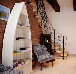 Una habitación con una estantería blanca curva, un sillón con estampado, una mesita de madera, un pequeño armario de madera y una escalera con barandilla de hierro forjado.