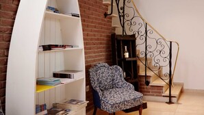 Una habitación con una estantería blanca curva, un sillón con estampado, una mesita de madera, un pequeño armario de madera y una escalera con barandilla de hierro forjado.