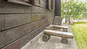 Una panca di legno rustica con base in cemento, appoggiata a un muro di legno consumato dal tempo con una finestra.