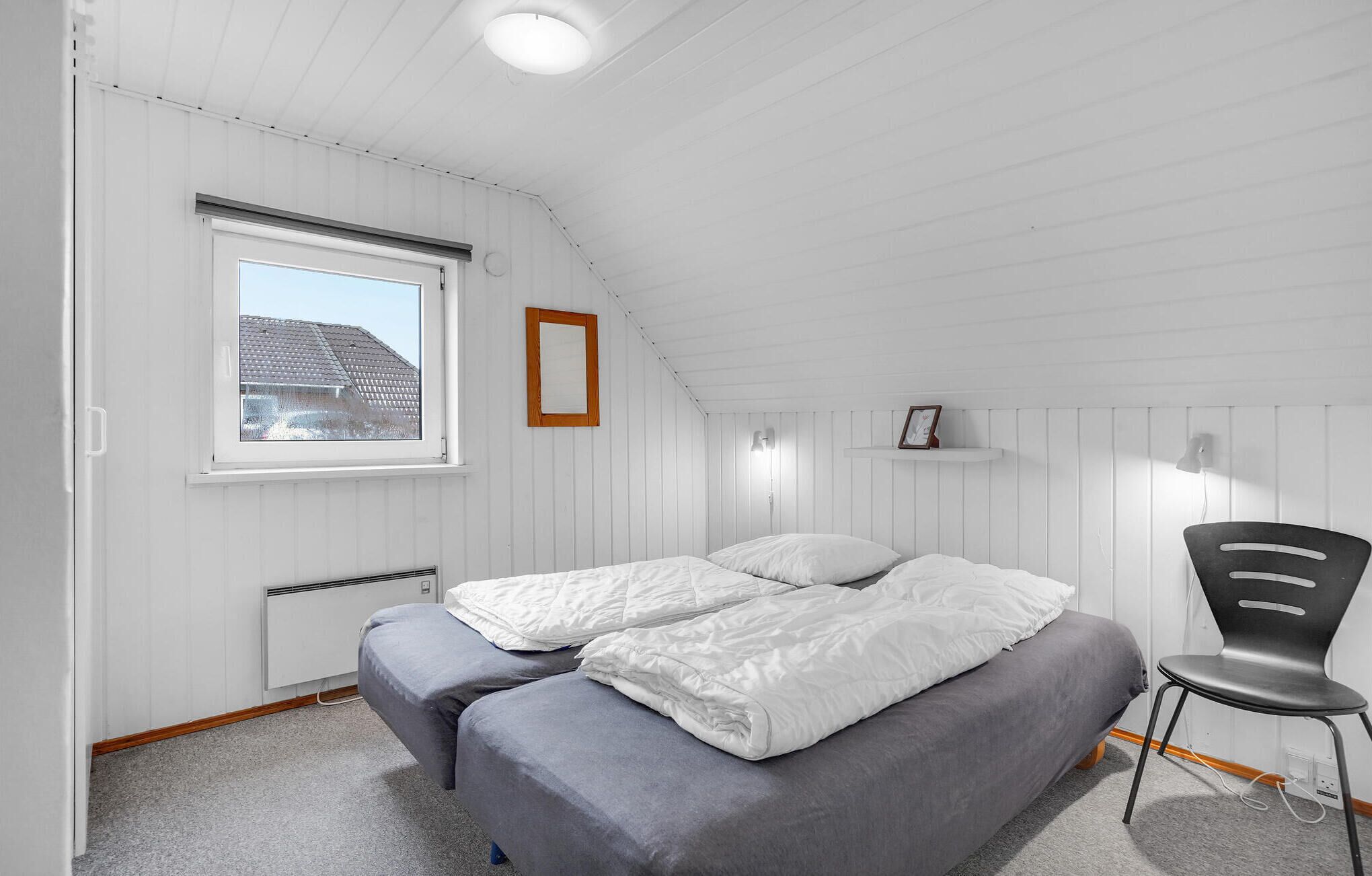 Una habitación pequeña con paneles blancos, una cama, una silla, un ventanal y un radiador.