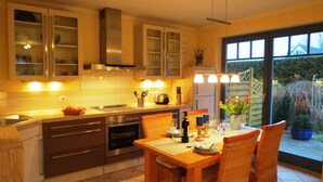 A modern kitchen with stainless steel appliances, a dining area with wicker chairs, and a view of a garden through a sliding glass door. - Gray Heron Apartment (Prerow)