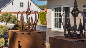 Two metal crown sculptures on concrete bases, with a house and greenery in the background.