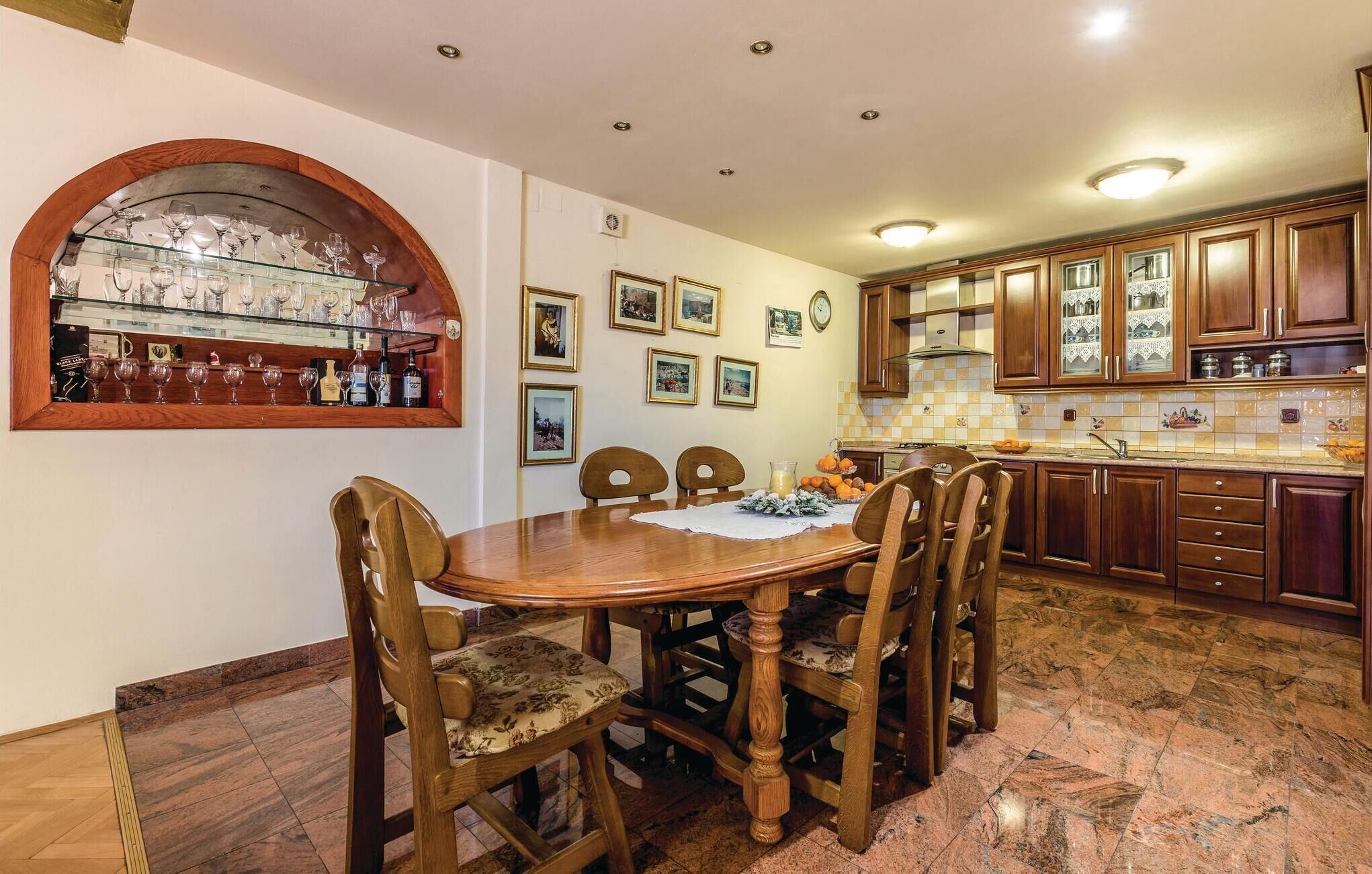 Un comedor con mesa y sillas de madera, una vitrina con copas de vino y una cocina con armarios de madera y salpicadero azulejado.