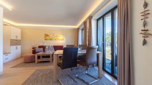Una habitación de hotel moderna con zona de comedor, sofá, mesa de centro y un amplio ventanal con cortinas.