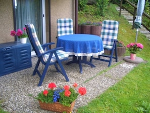 Un patio exterior con mesa y sillas, un pequeño jardín con flores y una escalera que lleva a un nivel superior.