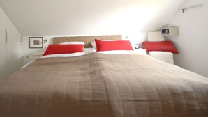 Une chambre à coucher avec un grand lit, deux oreillers rouges, une table de chevet avec une lampe et un réveil fixé au mur.