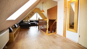 A spacious attic room with a sloped ceiling, wooden flooring, a black sofa, a wooden staircase, and a large window providing natural light. - Apartment 3 - Vacation Home Strandstr. 29 (Loddin)