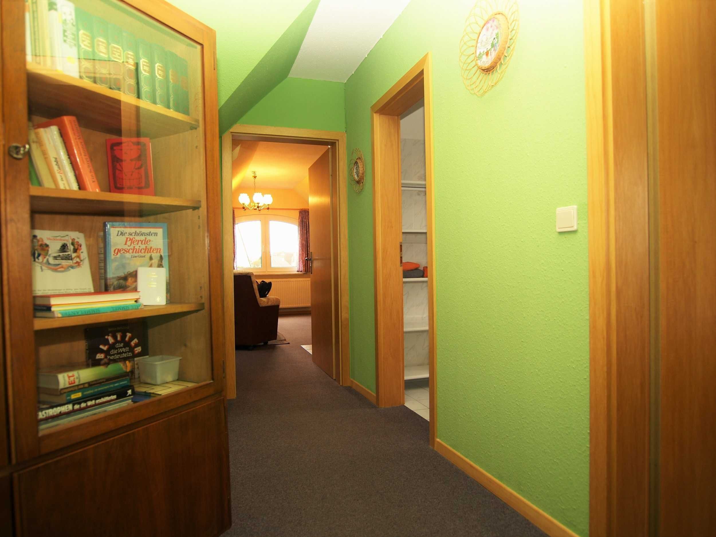 Un couloir aux murs verts, avec des portes en bois et une bibliothèque remplie de livres.