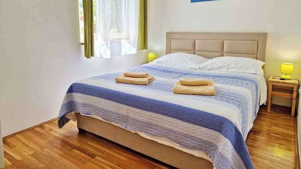 A neatly made bed with a striped blanket, two rolled towels, and a painting of a seascape on the wall. - Holiday flat with air conditioning and terrace (Baška)