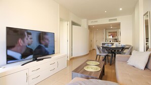 Een moderne woonkamer met een flatscreen televisie, een eethoek met een tafel en stoelen, en een bank met kussens.
