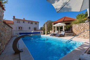 A swimming pool with lounge chairs and a gazebo, surrounded by stone walls and a building with a red-tiled roof. - Holiday apartment with shared pool (Crikvenica)