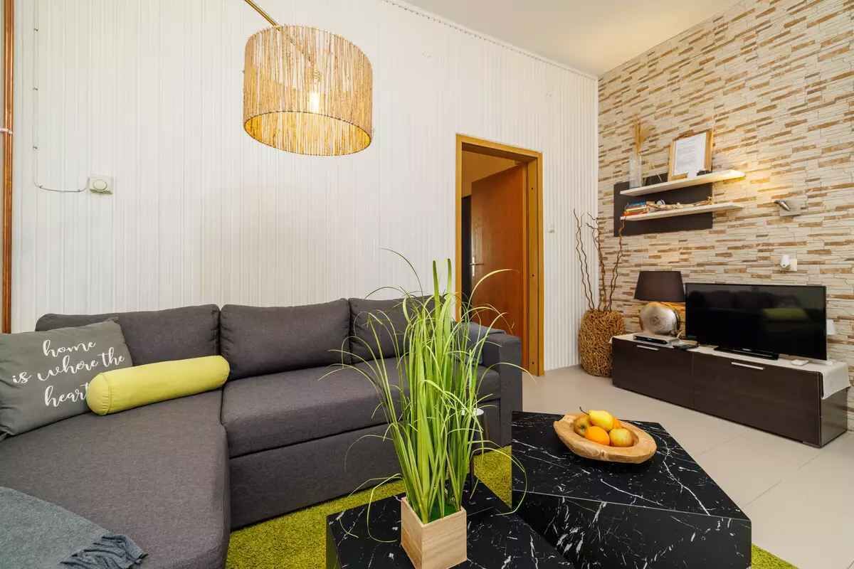 Un salón moderno con un sofá gris, una mesa de centro negra, una mesita auxiliar de madera con un cuenco de fruta, un televisor sobre un mueble negro, y una pared con un diseño tipo piedra.
