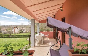A covered patio with a table and chairs, potted plants, and a view of a green landscape.