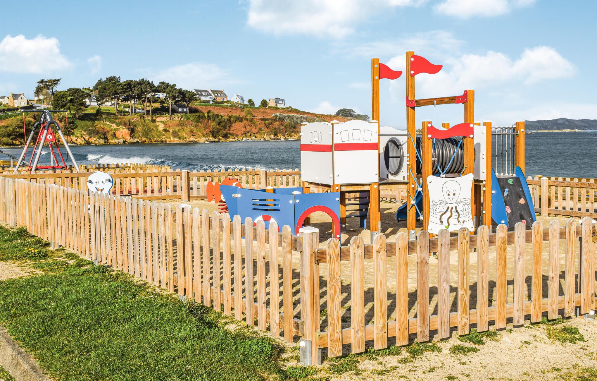 Une aire de jeux au bord de la plage, avec une clôture en bois, des toboggans et un poste de surveillance pour les sauveteurs.