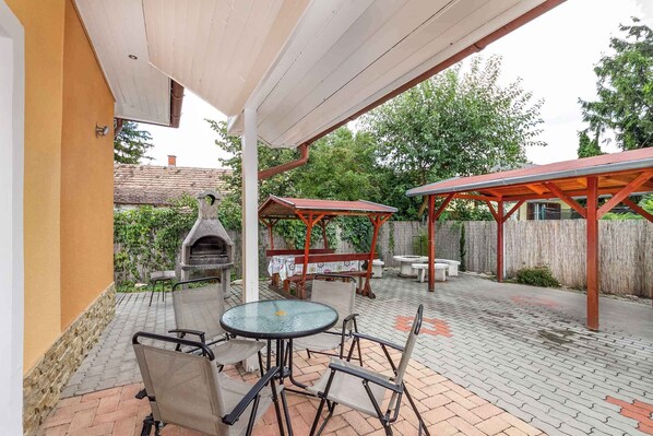 Un patio al aire libre con piso de ladrillo, sillas de metal, una mesa de vidrio, una parrilla y una pérgola de madera.