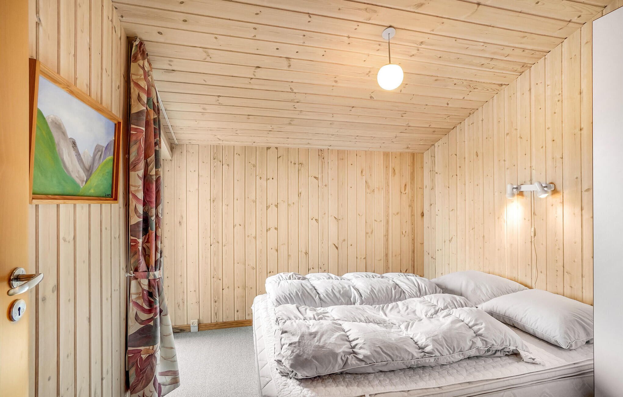 Ein Zimmer mit Holzwandverkleidung, einem Bett mit gesteppter Matratze und Kissen, einem Gemälde an der Wand und einer Deckenleuchte.