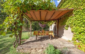 Un espace extérieur aménagé avec des sièges, protégé par un toit, doté d’un sol en pierre, entouré de verdure et bordé d’un mur en pierre.