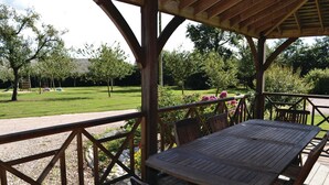 Uma área de estar coberta ao ar livre, com mesa e cadeiras, rodeada por um deck de madeira e um jardim com diversas plantas e flores.