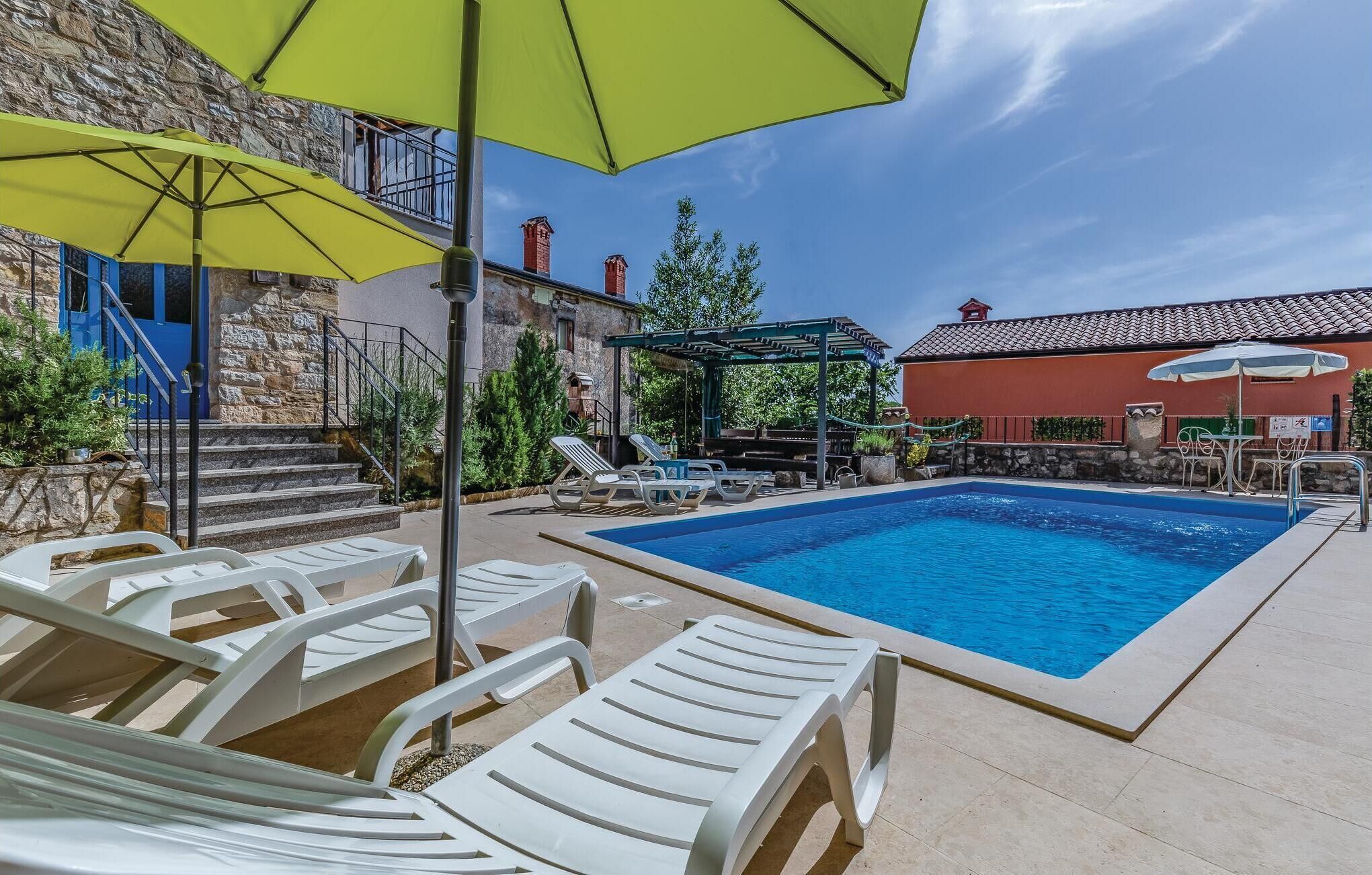 A swimming pool with lounge chairs and umbrellas, a stone building, and a red-roofed structure.