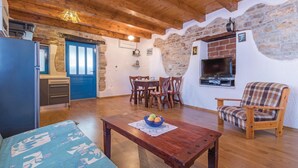 A cozy living room with a wooden ceiling, stone walls, a dining area with chairs and a table, a sofa, and a TV mounted on the wall.