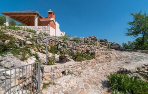 Situated on a sloped terrain, this traditional building features a metal pergola, stone wall, and garden area with lush plants and flowers.