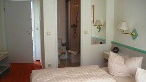 A hotel room with a bed, a nightstand, a mirror, a wall-mounted lamp, and a shower area. - Double room small - Hotel Reke (Hotel-Admin) (Plau am See)