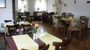 A dining area with wooden tables and chairs, a vase with flowers, and a view of the dining room through a doorway. - Double room small - Hotel Reke (Hotel-Admin) (Plau am See)