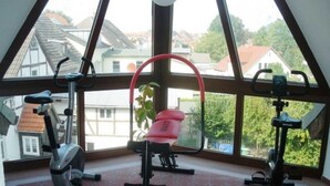A gym with exercise equipment and a large window offering a view of houses and trees. - Double room small - Hotel Reke (Hotel-Admin) (Plau am See)