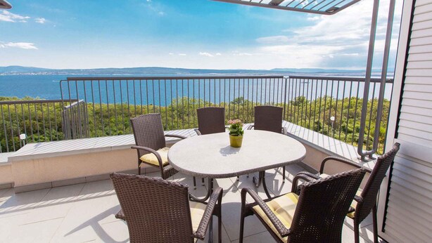Een balkon met een ronde tafel en stoelen, een plant in pot en uitzicht op zee en groen.