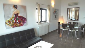 Een moderne woonkamer met een zwarte leren bank, een eettafel met stoelen, een groot schilderij aan de muur en een raam met gordijnen.