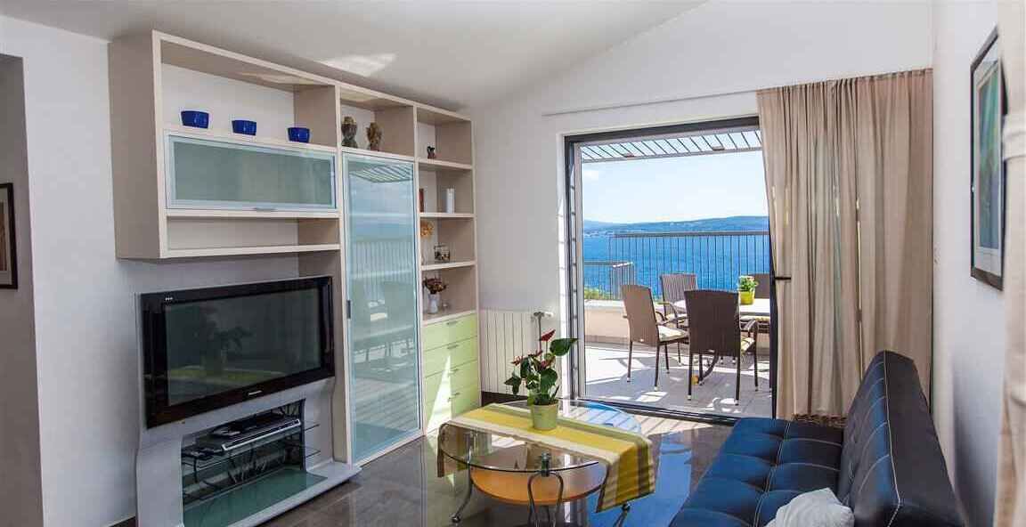 A modern living room with a flat-screen TV, a sofa, a dining table with chairs, and a balcony with a view of the sea.