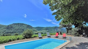 A rectangular pool with clear blue water, surrounded by a gravel area. There are lounge chairs, some red and some grey, and a large tree providing shade. The background features a hilly landscape with greenery. - Amazing home in Flaviac (Flaviac)