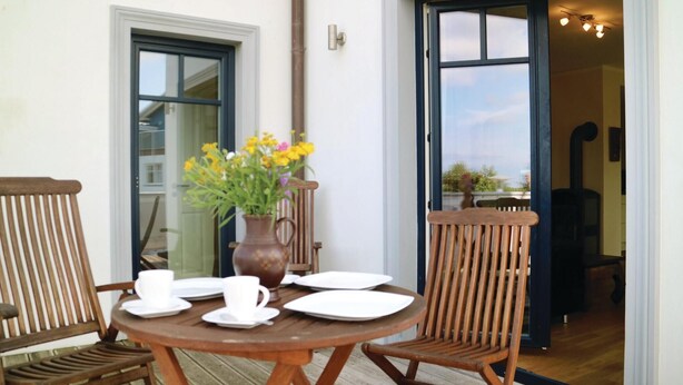 A wooden table set for two with white plates and cups, a vase with yellow flowers, and two wooden chairs.