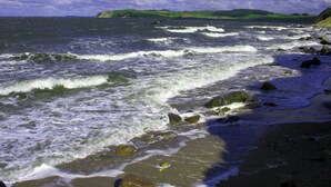 Ein Strand mit Wellen, Felsen und einer entfernten grünen Hügelkette.