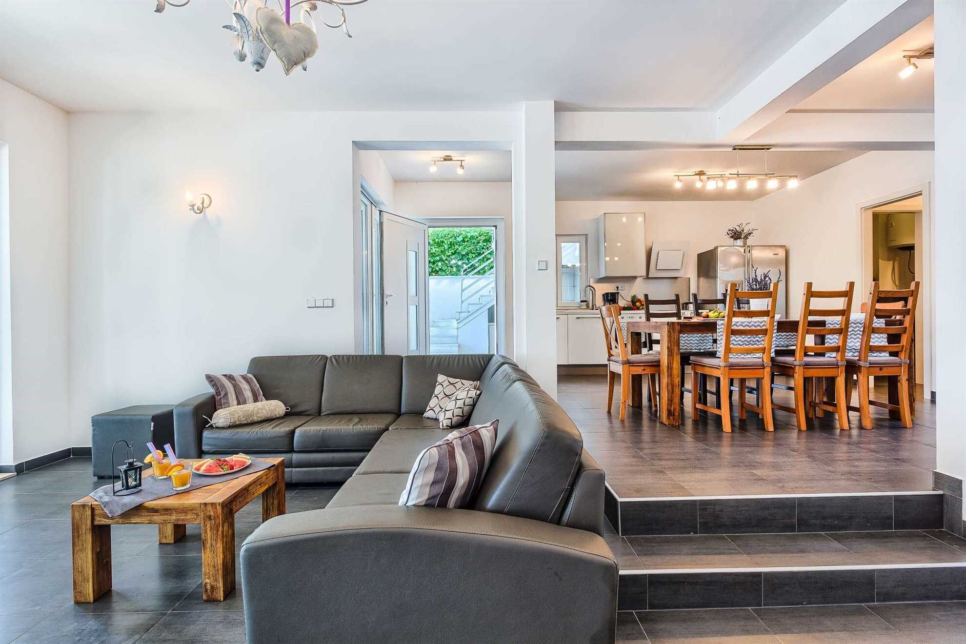 Un salon moderne avec un canapé d’angle, une table basse en bois sur laquelle sont disposés des fruits et des boissons, et un coin repas avec des chaises et une table en bois.